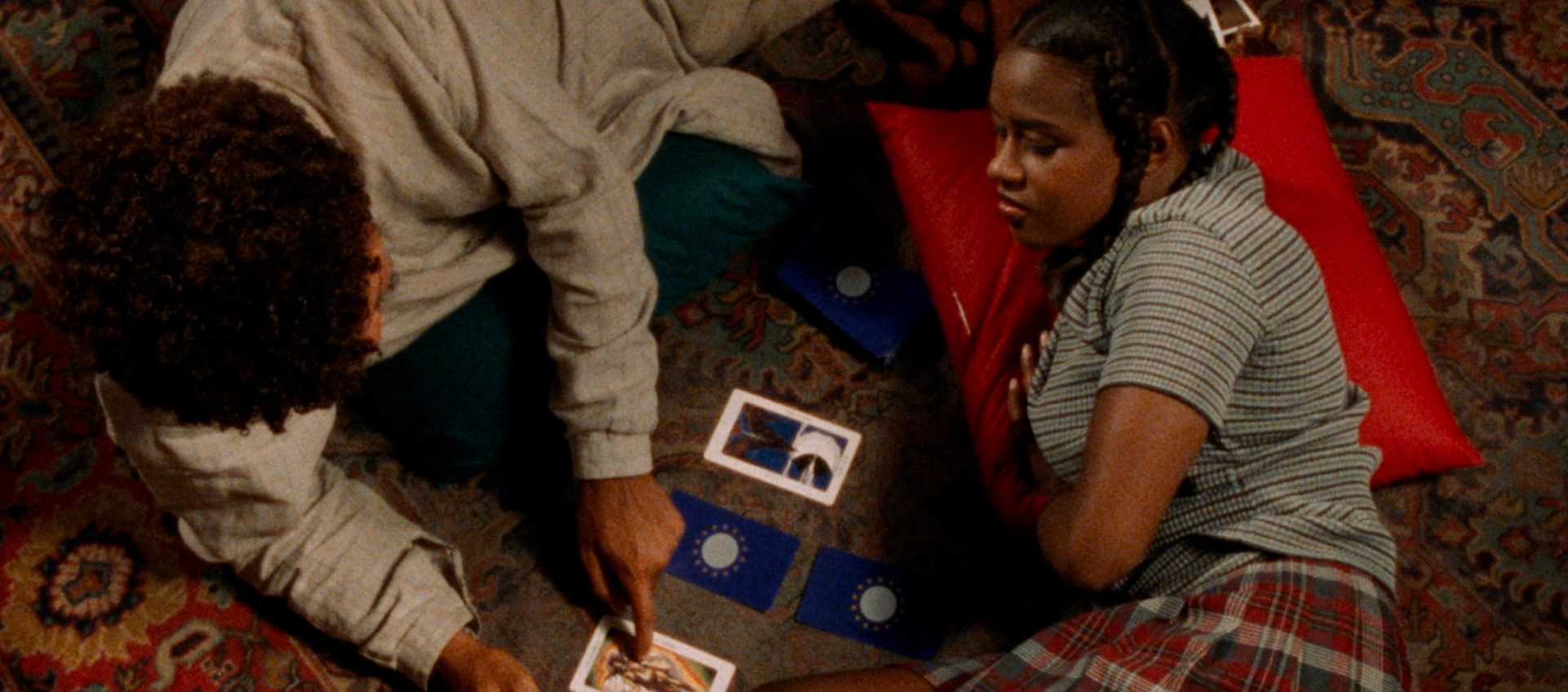 Overhead view of a young Black man and a young Black woman lying on a decorative carpet. He’s pointing to one of four tarot cards laid out between them.