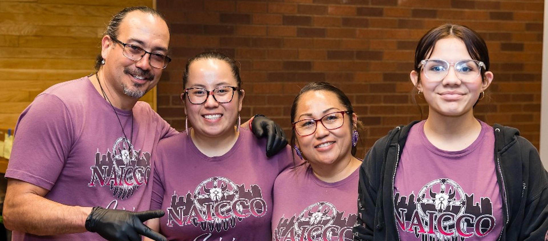 A lineup of one man and three women of Native American descent. All are wearing light purple T-shirts with the black and gold logo for NAICCO Cuisine.