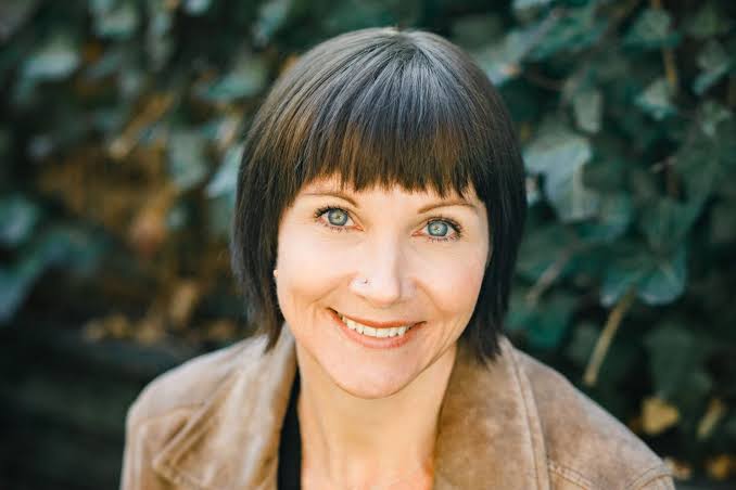 A woman with straight, short, brunette hair and a beige jacket. She’s sitting outside with a leafy background and she’s smiling.