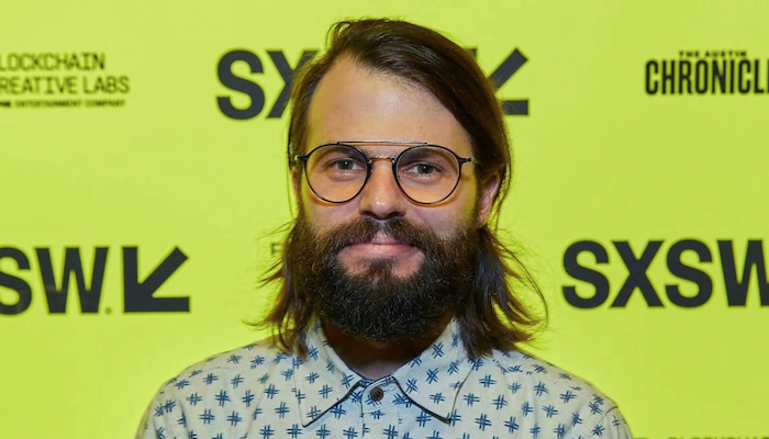 A color photo of Colin West, a white man with dark brown shoulder-length hair and facial hair, in front of a bright yellow background with the SXSW logo on it.