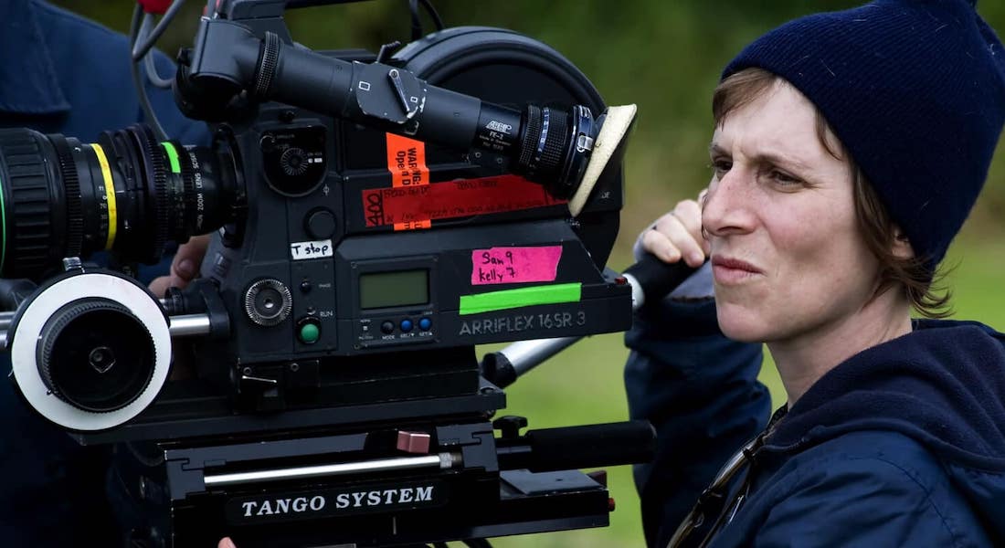 Filmmaker Kelly Reichardt looks through the viewfinder of a 16mm camera during a shoot outdoors. She is a white woman with short brown hair, wearing a navy beanie cap and hooded jacket.