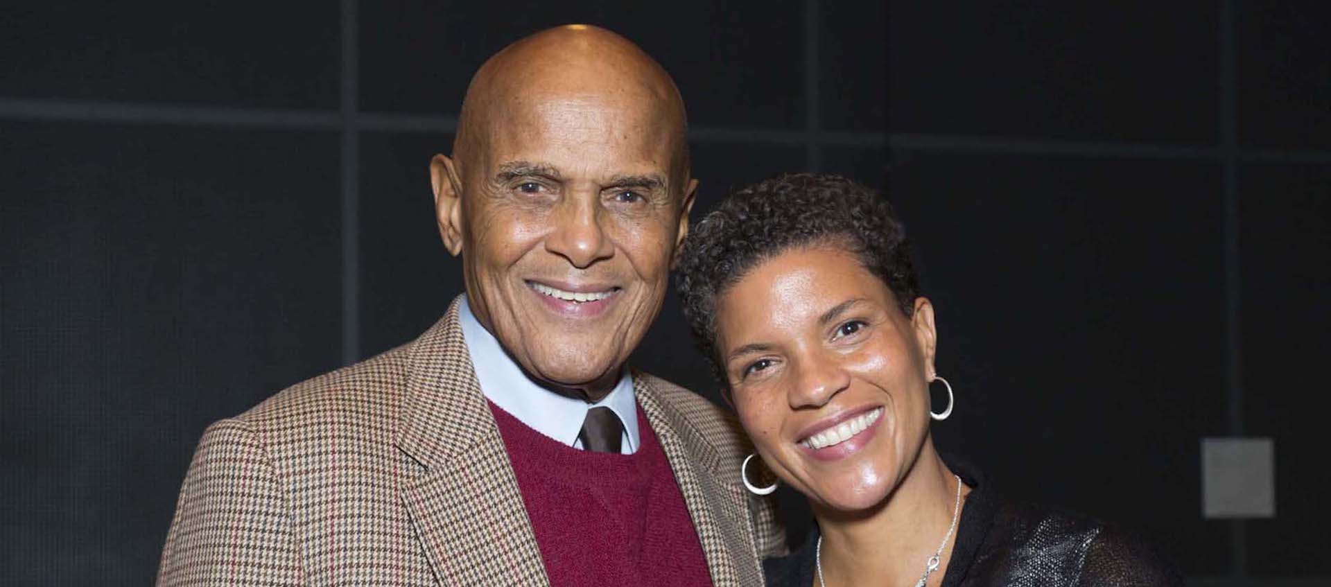 An older, bald man in a suit jacket, tie, and sweater smiles for the camera with a younger woman by his side, also smiling as she leans close to him.