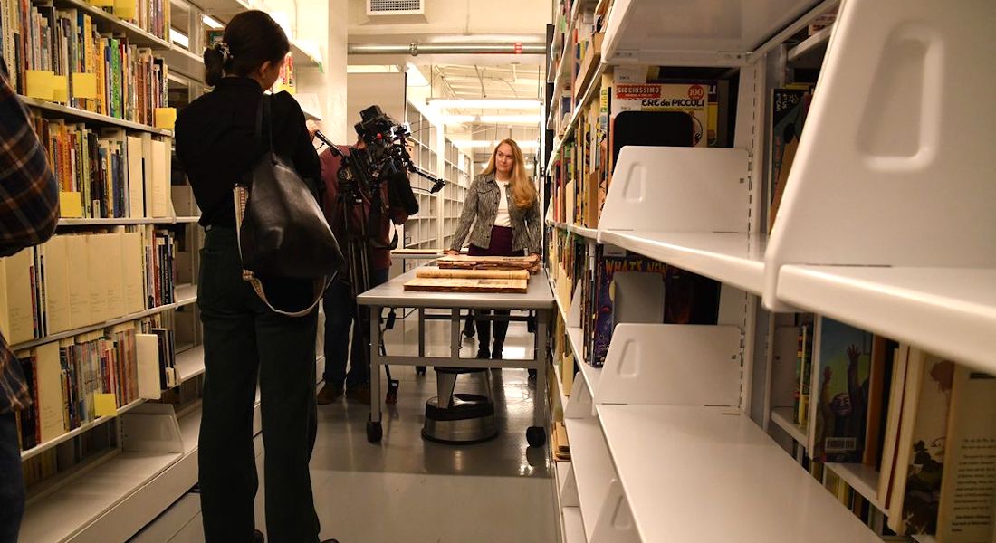 A woman stands at the end of a long table in a library corridor with tall shelves lining either side. A camera operator captures footage of her while another woman looks on in the background.