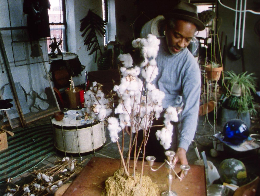 A man in a rimmed hat and sweatshirt stands in a plant and object-filled studio. In front of him on a table is a sculptural work that features branches from an unpicked cotton plant.