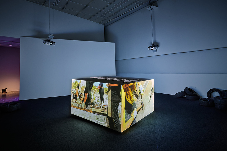 A cube in the center of a dimly lit gallery space casts video projections. There are tires placed on the floor to the right.