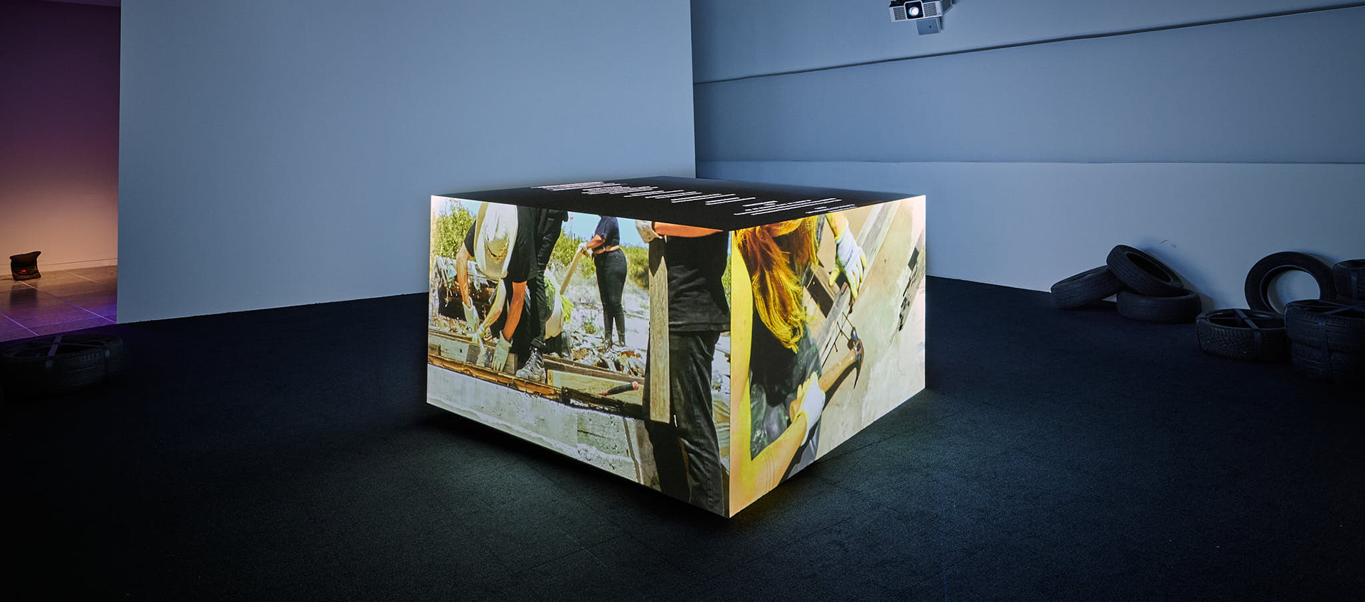 A cube in the center of a dimly lit gallery space casts video projections. There are tires placed on the floor to the right.