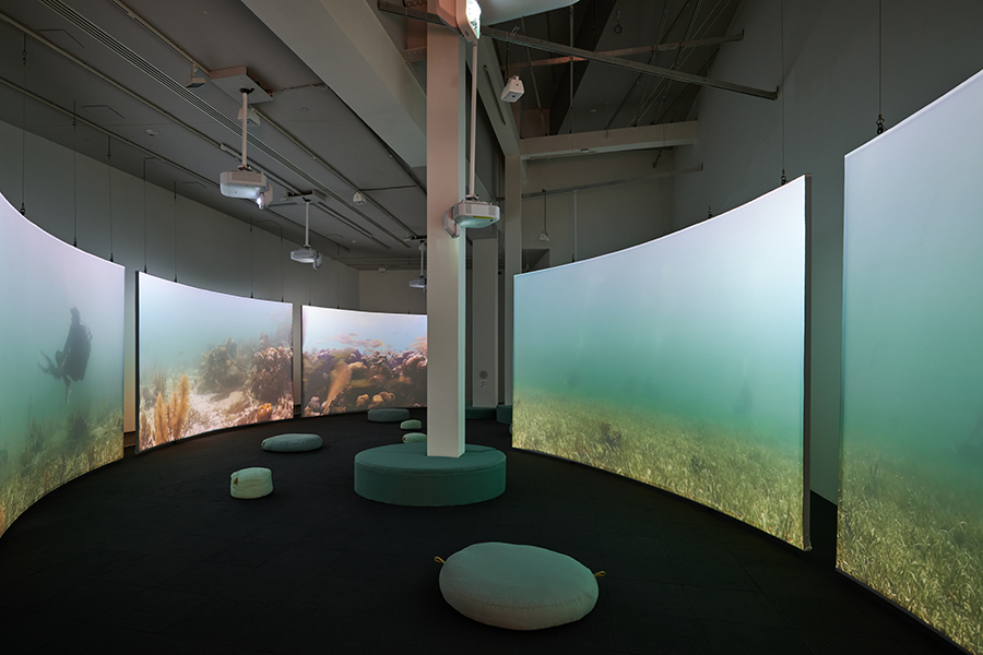 A dimly lit gallery space surrounded by five large, curved screens projecting scuba divers meditating on the ocean floor. Round cushions are distributed throughout the space.