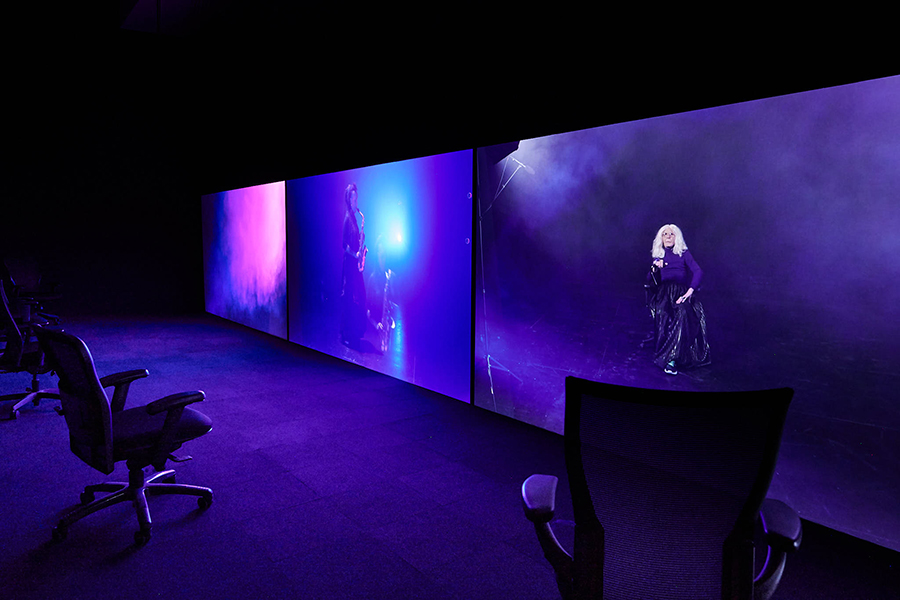 A video installation in a dimly lit space with three large screens emitting pink and purple hues. There are three chairs placed in front of the screens.