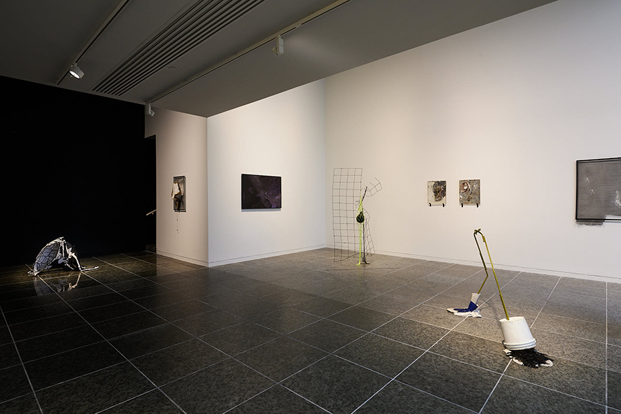 A gallery space lit by overhead lights has three sculptural objects spaced throughout the black tile floor. Five works hang on three gallery walls.