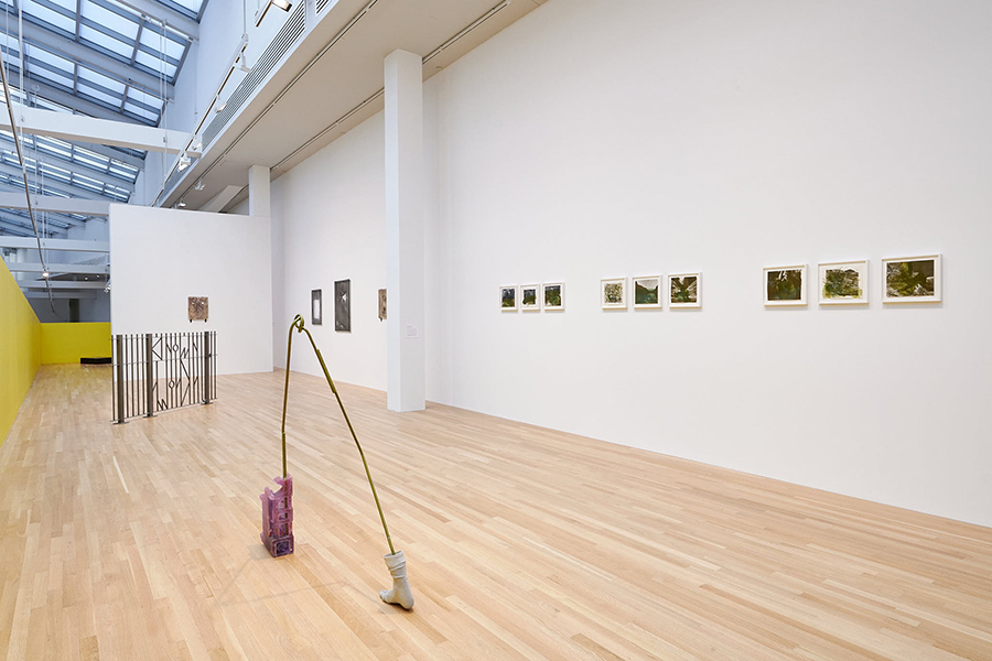In a long gallery space, two sculptures are placed on a wooden floor, and artworks are hung on the right and back wall. A yellow wall emerges near the end of the gallery.
