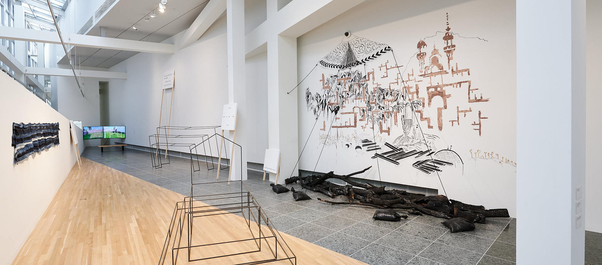Metal sculptures and burnt tree branches are on the floor of a gallery space featuring white beams. Works are painted and hung on the walls. A video plays in the background.