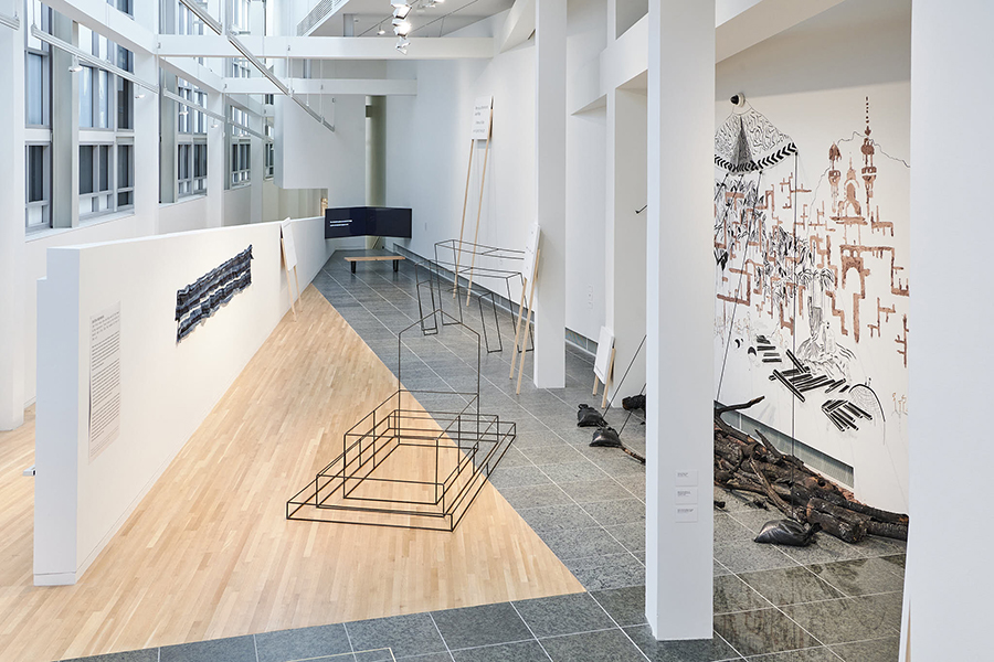 A gallery space viewed from above features metal sculptures on the floor, a mural on the right wall, hung denim textile on the left wall, and signs leaning against white beams.