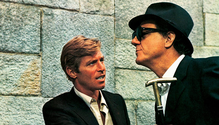 Two men wearing dark suits are facing each other in front of a stone wall. One is looking at the other, who is wearing a dark hat and glasses and carrying a cane. 