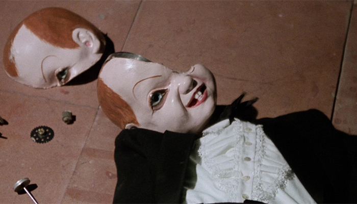 A ventriloquist dummy with wearing a black suit is lying on the ground. Its head is shattered with the right side resting above the body.