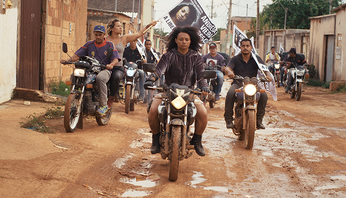 A group of Brazilians ride mud-covered motorcycles down a muddy favela street, led by a woman and carry flags with her face and Portuguese text on them. 
