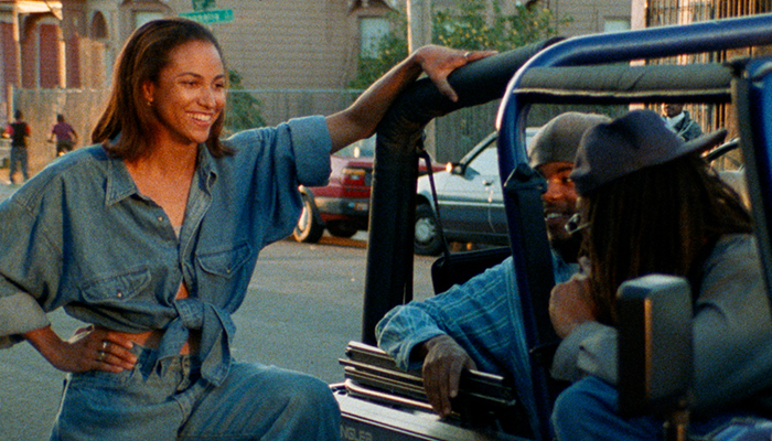 Tobi (April Barnett), a young Black woman wearing a denim outfit, stands on the side of a Jeep talking to two Black men seated inside the vehicle.