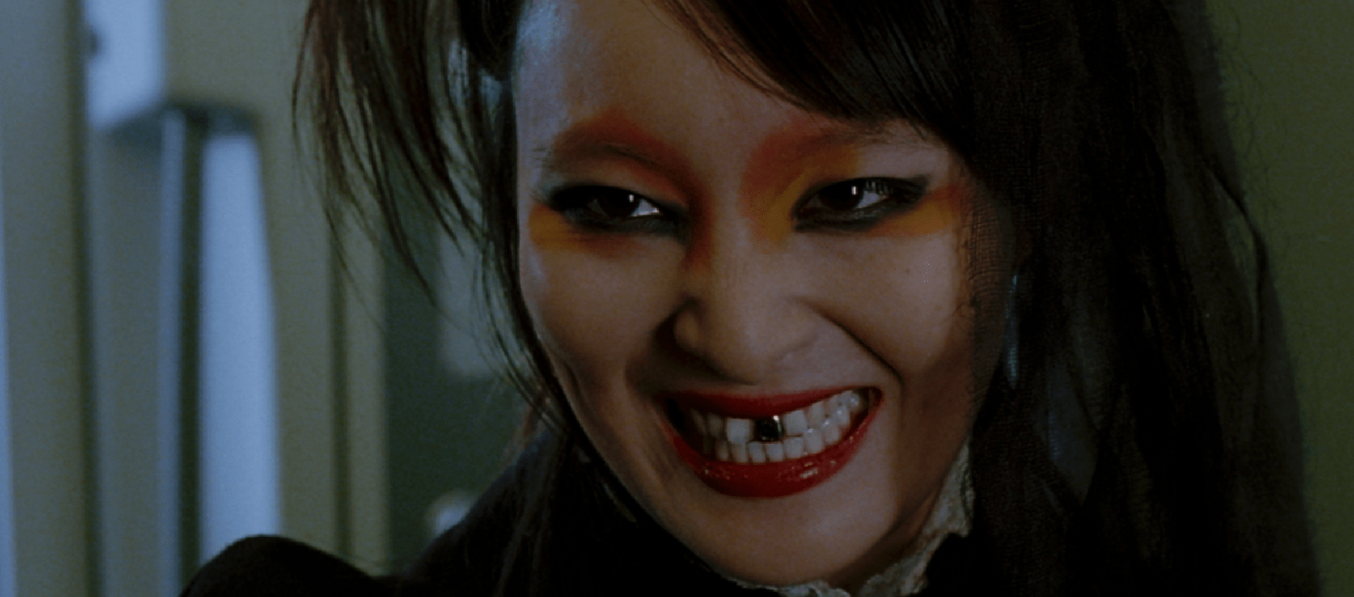 A close-up of a woman with an evil grin. She has dark hair and dark eye makeup. One of her front teeth is made of gold. 