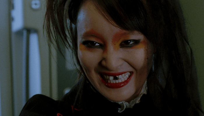 A close-up of a woman with an evil grin. She has dark hair and dark eye makeup. One of her front teeth is made of gold. 