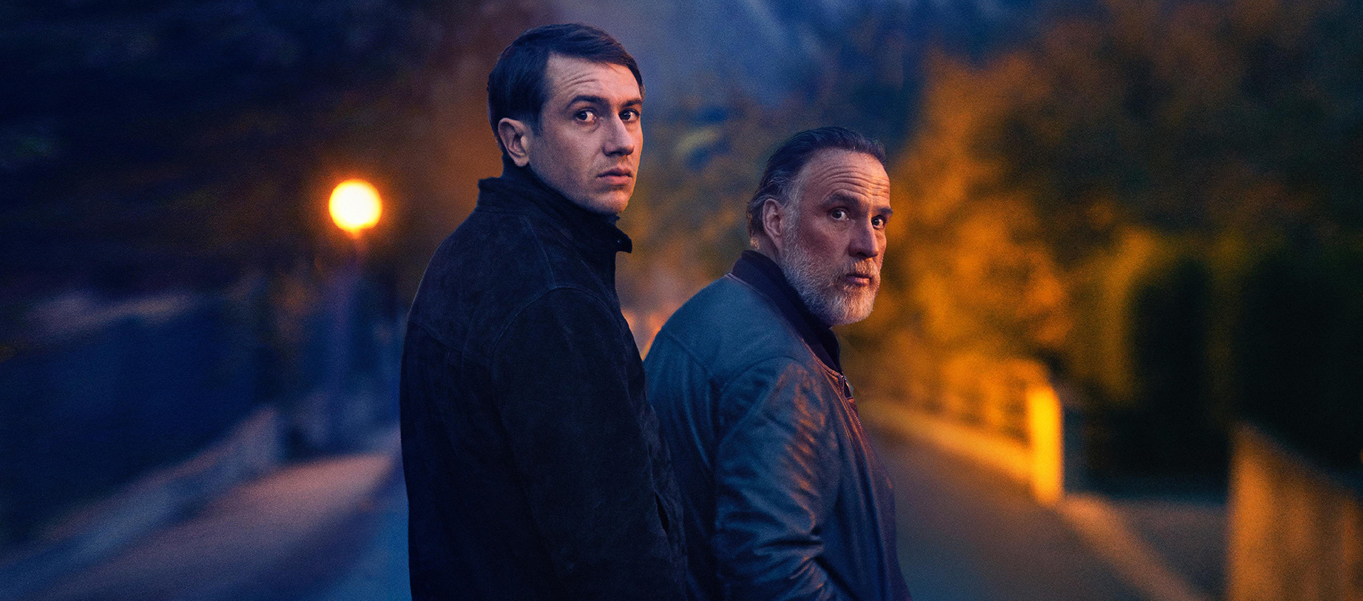 Two men, one tall and one shorter with a gray beard, are standing in the middle of the street at night, turning back to look over their shoulders. 