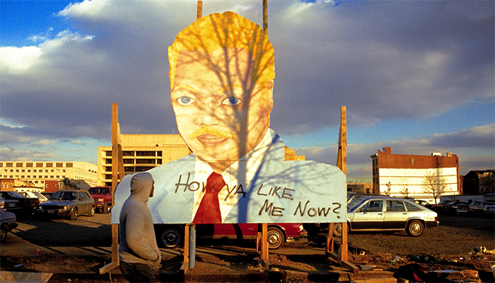 A man looks at a billboard, painted by David Hammons, that depicts Reverend Jesse Jackson with pale skin, blonde hair, and blue eyes. “How Ya Like Me Now?” is painted across Jackson’s blue suit.  