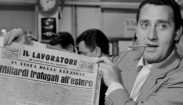 Black-and-white film still featuring Silvio (Italian actor Alberto Sordi), who has a pencil in his mouth and holds up an Italian newspaper.