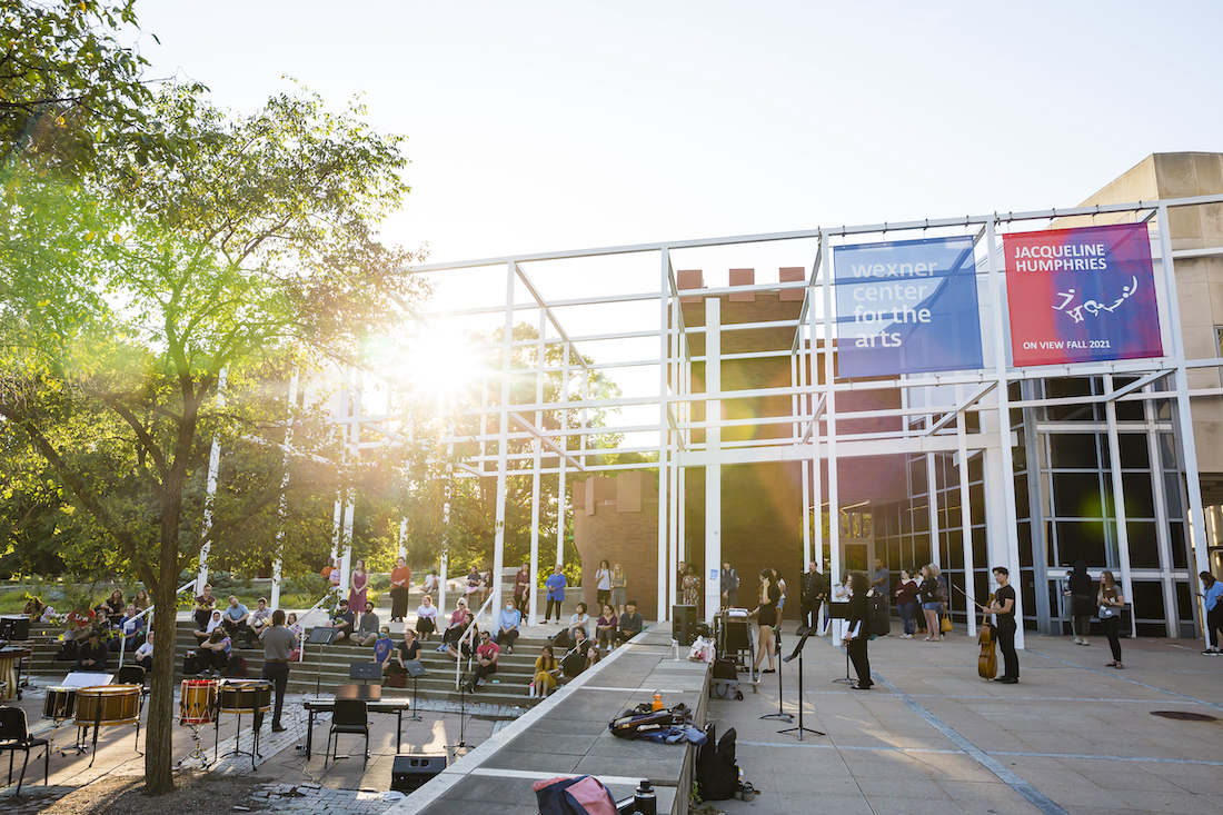 An outdoor plaza filled with people watching a music performance. The plaza is adjacent to an arts center building in a postmodern style with a white metal grid around its exterior. A banner attached to the grid identifies the building as the Wexner Center for the Arts. The sun is setting in the background.