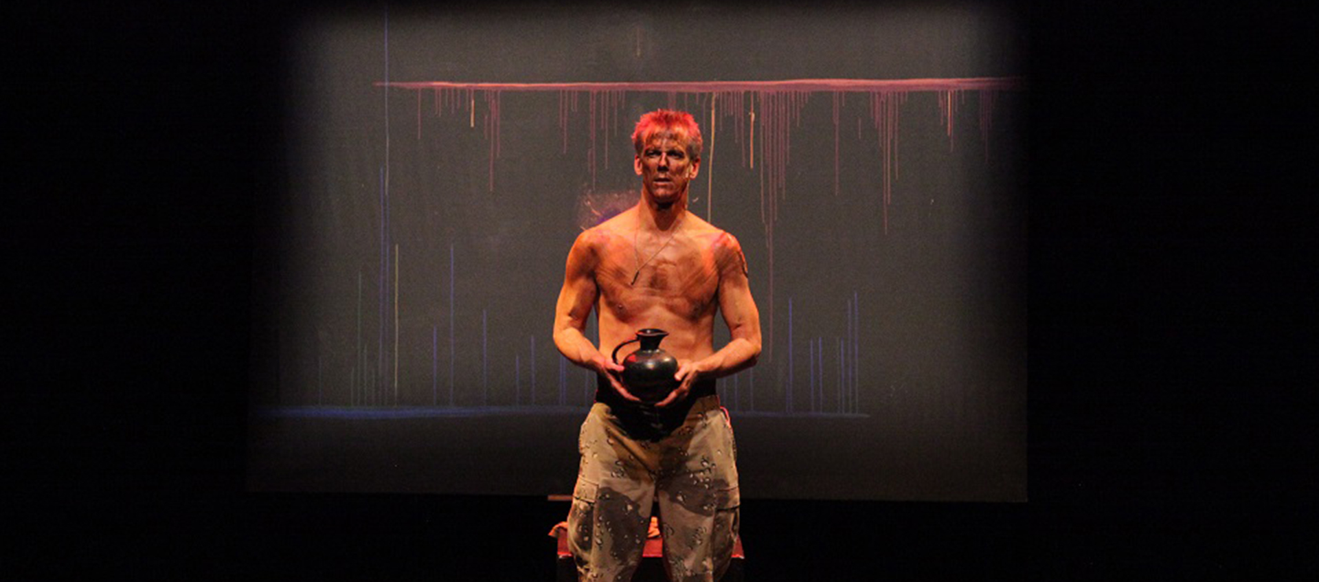 A tattered, shirtless man in camouflage pants holds a black vase on a red lit stage.