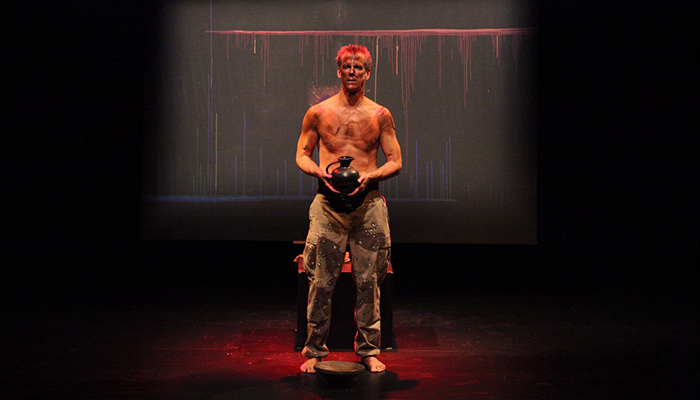 A tattered, shirtless man in camouflage pants holds a black vase on a red lit stage