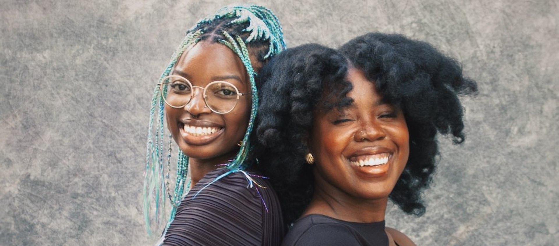 Two young black women standing with their backs against each other, each with a big, warm smile.