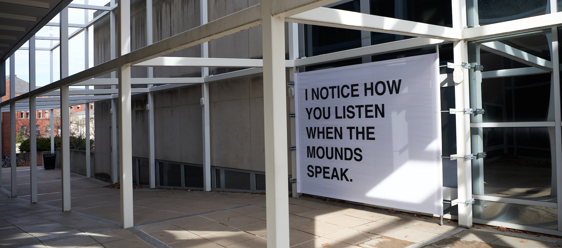 A white sign with black text is installed outside against an architectural grid framework. 