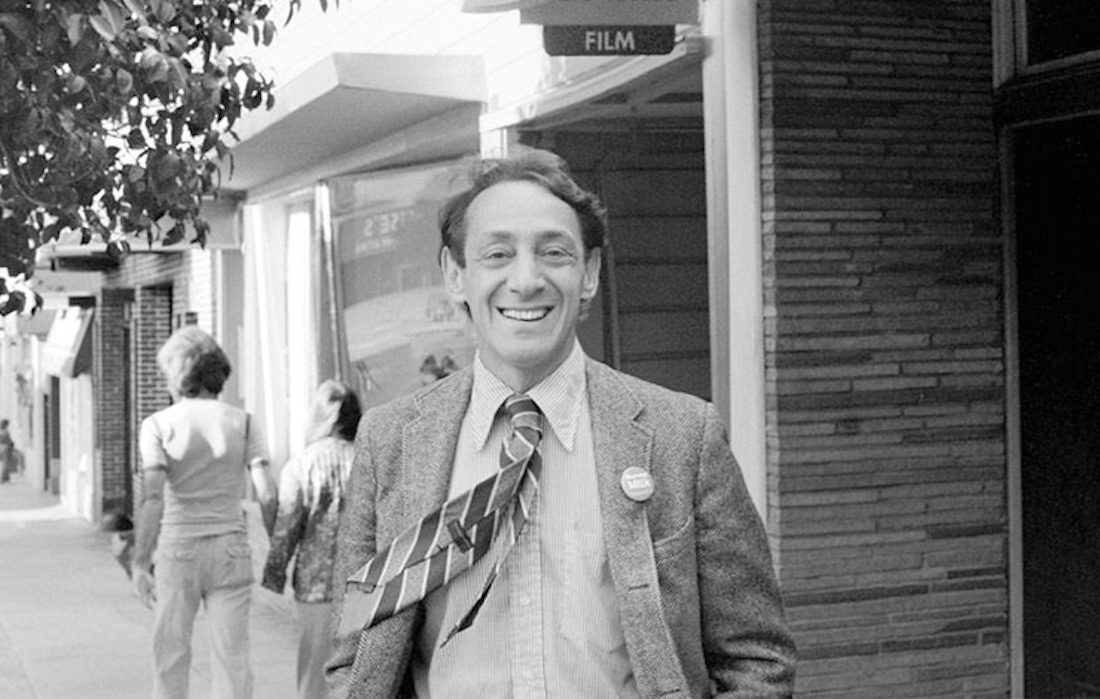 Black and white image of a man on the street. He has dark hair and is in a jacket and tie, and he’s smiling for the camera