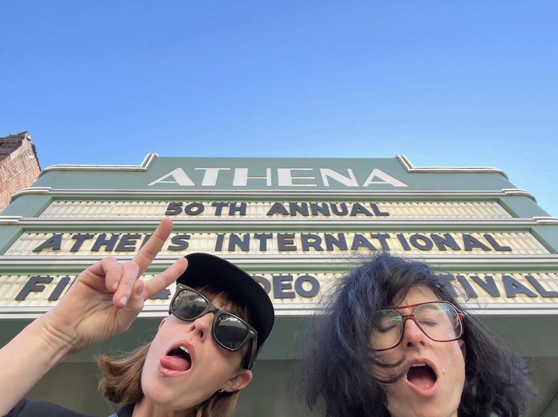 A low-angle shot of two women making slightly funny faces. A theater marquee and a blue sky is behind them