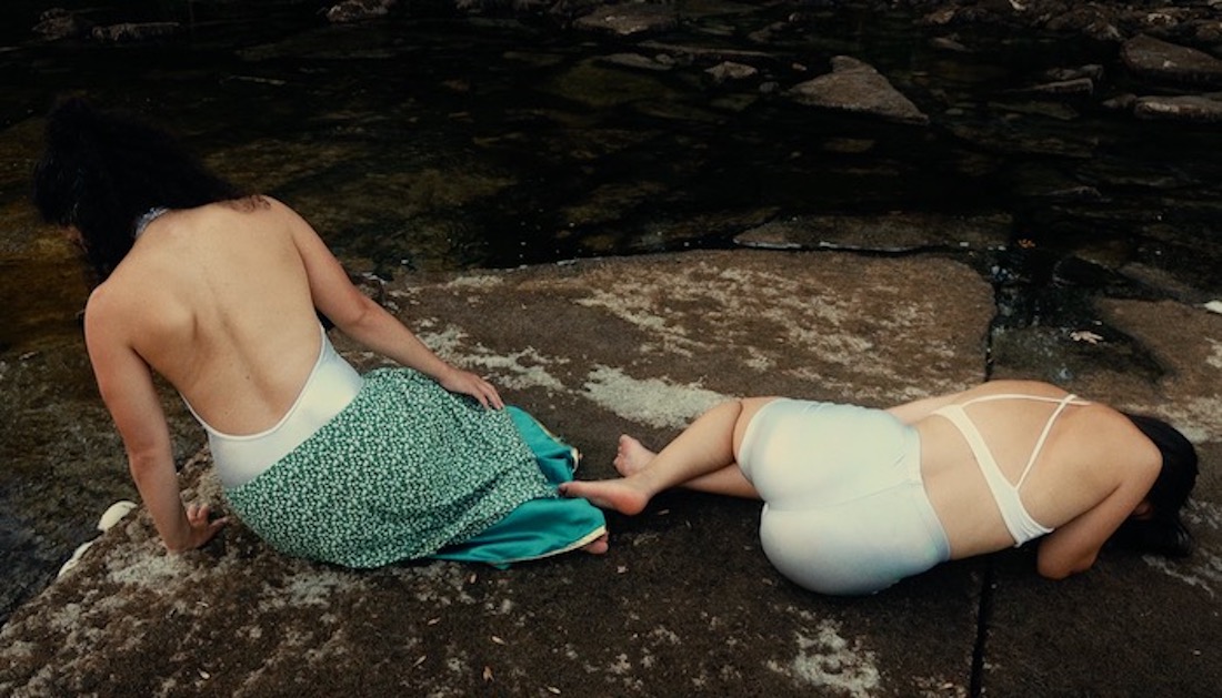 A video still of two women wearing swimsuits, facing away from the camera. They are outdoors and stretched out on a large, flat rock.