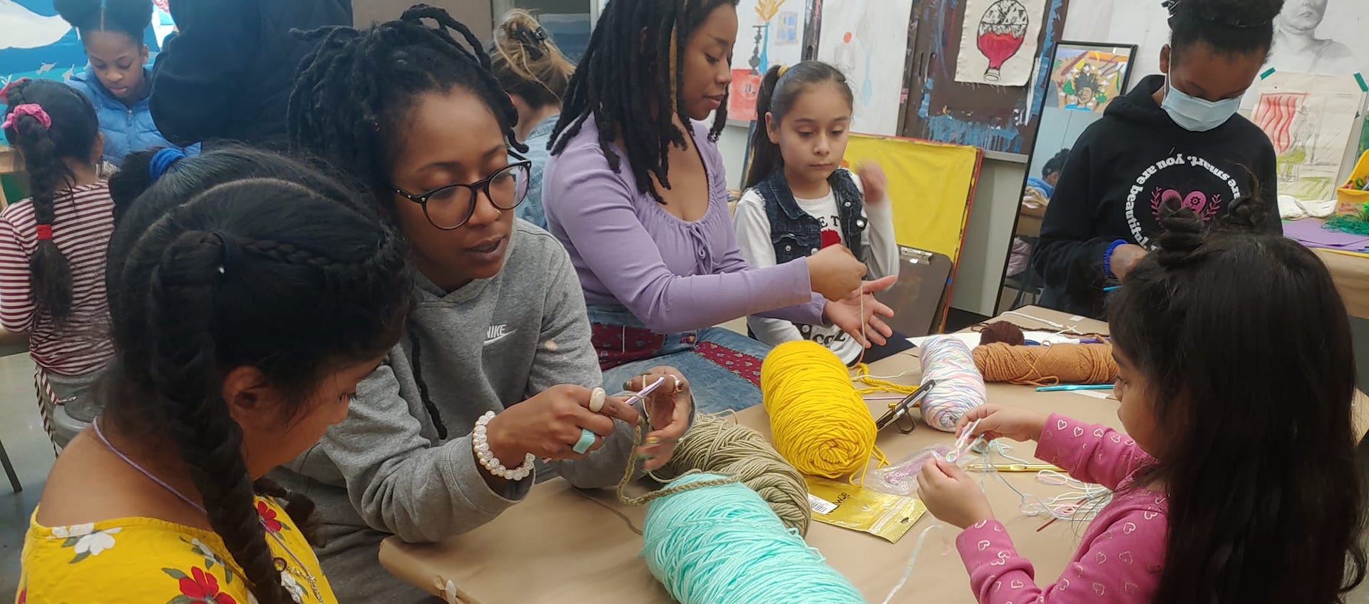 A group of six women and girls sit together in a classroom at a long table. Each is knitting from skeins of yarn placed on the table. Student artworks are covering the wall behind them.