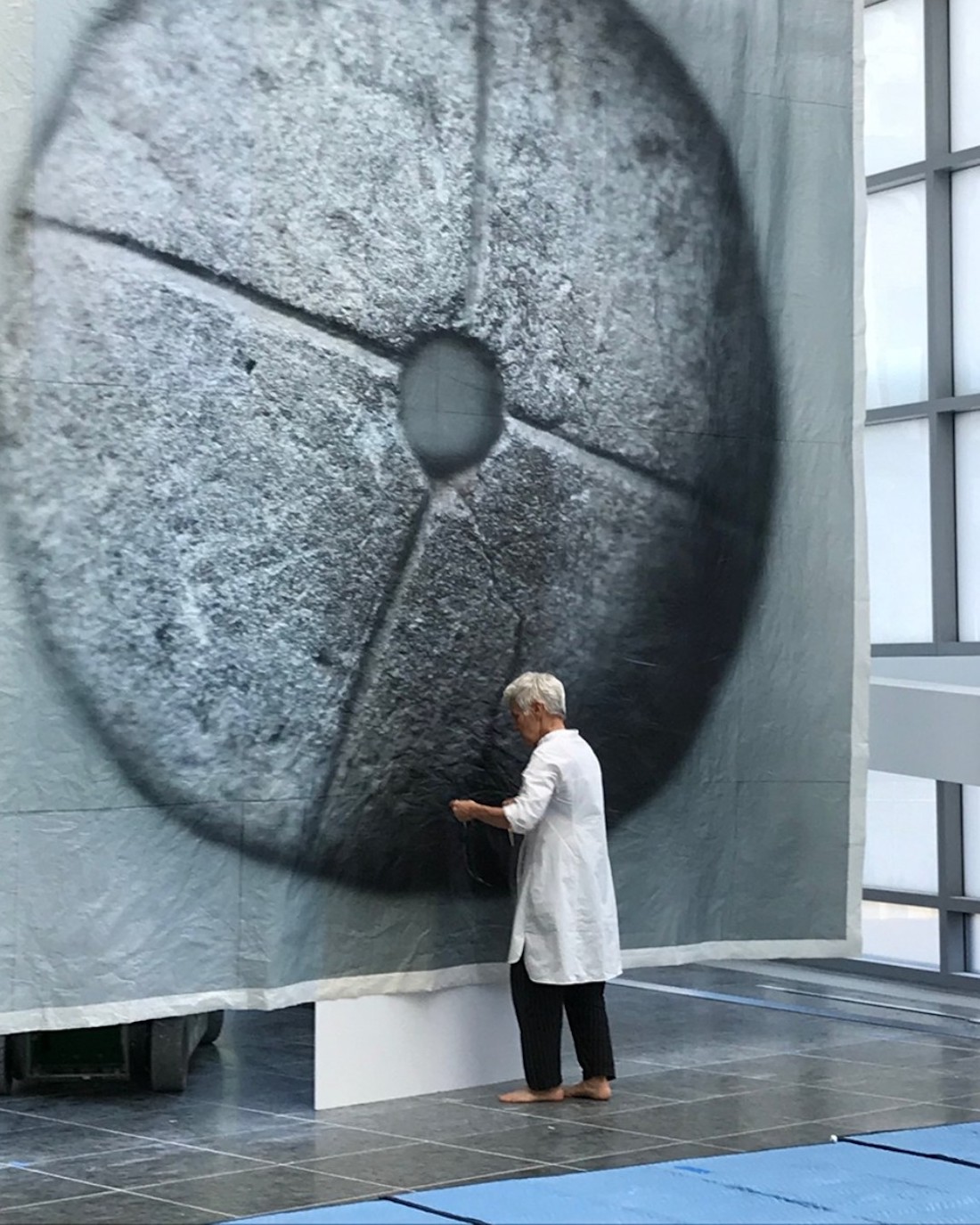 A woman with short white hair, in a long white tunic and black pants, stands in a large, high-ceilinged gallery space. Directly in front of her is a towering wall hanging holding an image scan of a flat stone wheel.