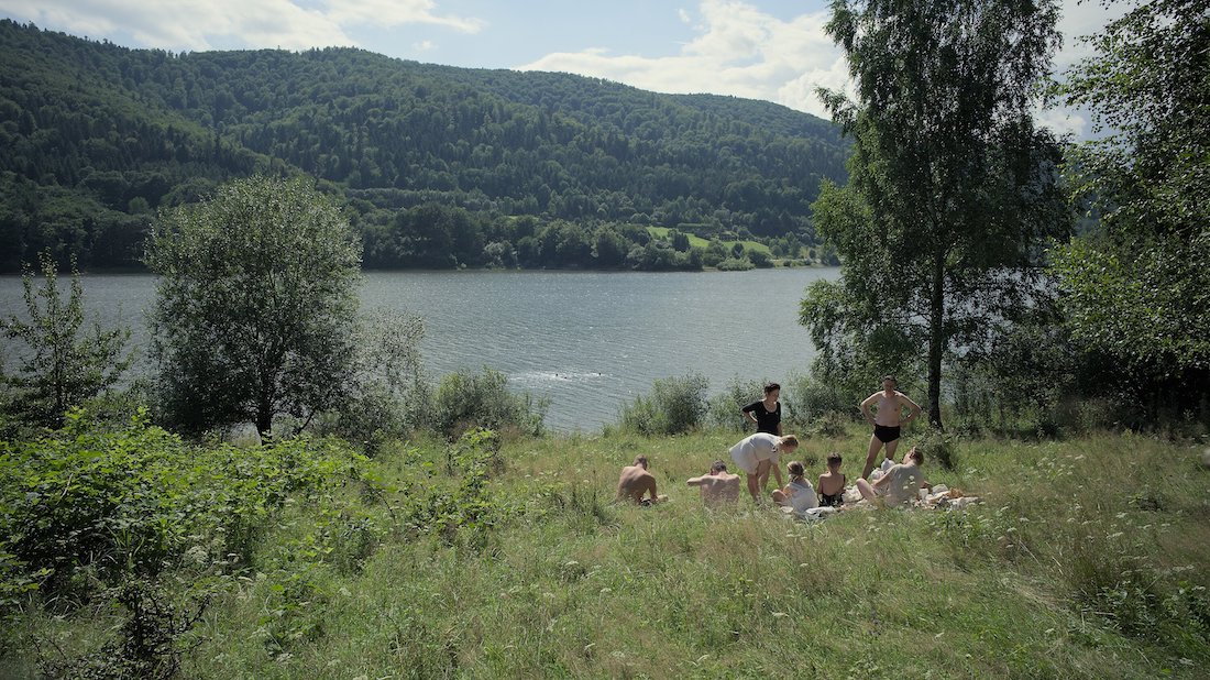 A wide shot of the grassy, tree lined bank of a body of water. A group of men and women in light clothing or swimsuits stand or sunbathe in the grass.