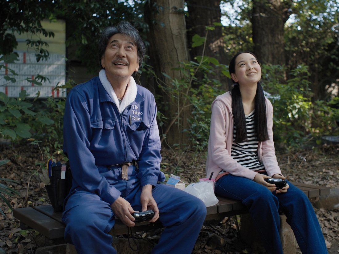 An older man and young woman sit in a forest, looking up at the sky and smiling.