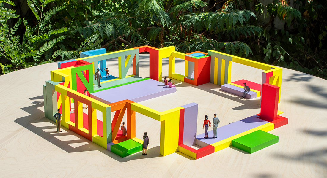 Overhead of colorful architectural mock-up. It is square in shape with a courtyard. The design elements are geometric in shape. Miniature figurines inhabit the space.