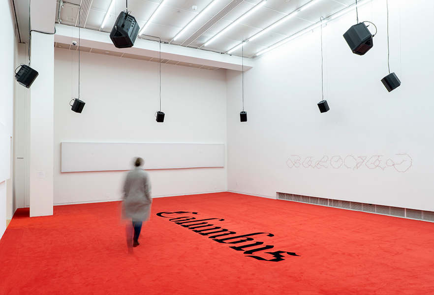 A circle of hanging speakers, a person in motion walks on red carpet with black text reading "Columbus." Text mural on wall reads "beloved."