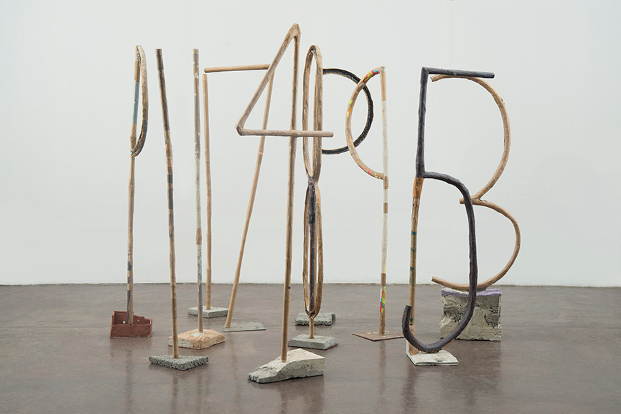 A cluster of tall and narrow sculptures of numbers with unique bases are placed on the floor in front of a white wall.