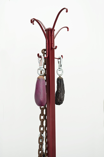 Two dried eggplant sculptures hang on two rungs of a deep cherry-red hat rack. A bulky chain is hanging on the left side of the rack.