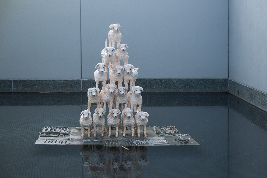 Sculpture with 15 small, white ceramic dogs stacked in a pyramid sits on a cement plinth in a pool. The sculpture's reflection is visible in the water. 
