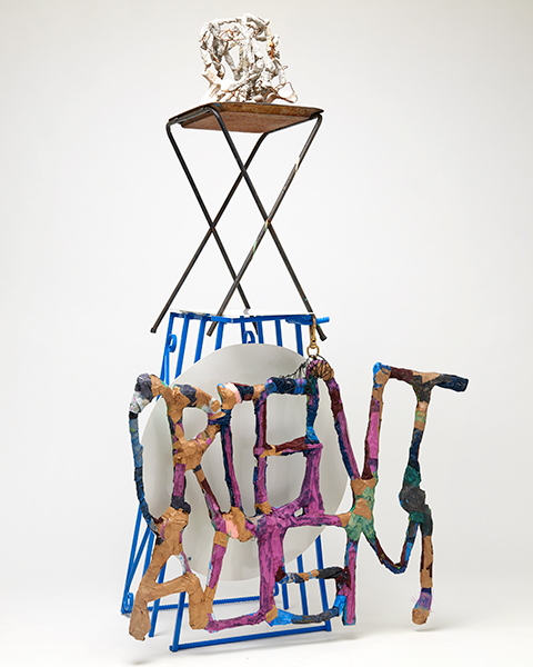 A sculpture with a blue, cage-like base supports a TV tray with a sculpture on top. The multicolored word Orientalism hangs like a charm in the front.
