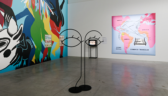 A vertical sculpture with eyes rests in foreground. A colorful mural is on the left. A small tv and large painting are on the right.
