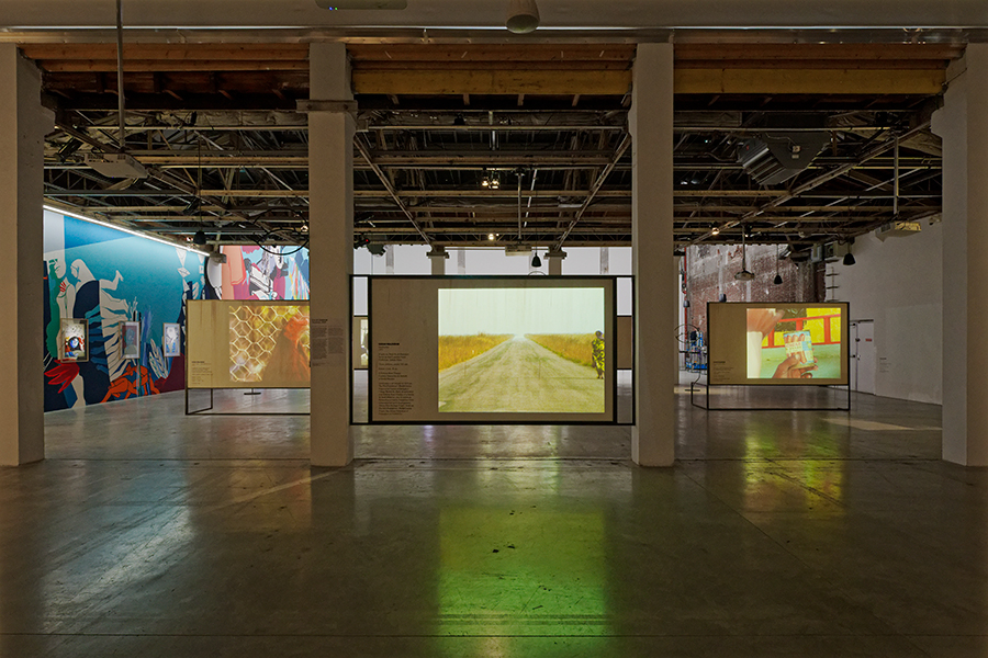 View of an exhibition with three films projected onto rectangular apparatuses. A colorful wall mural to the left extends backward.