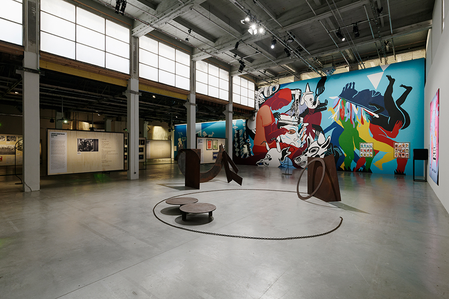 Installation view of a multi-piece metal sculpture made up of multiple shapes with a colorful wall mural in the ear.