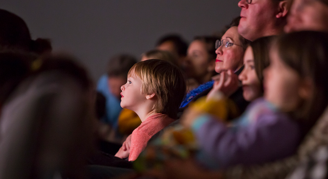 A close-up of a family seated in a theater with the smallest child looking up at the screen