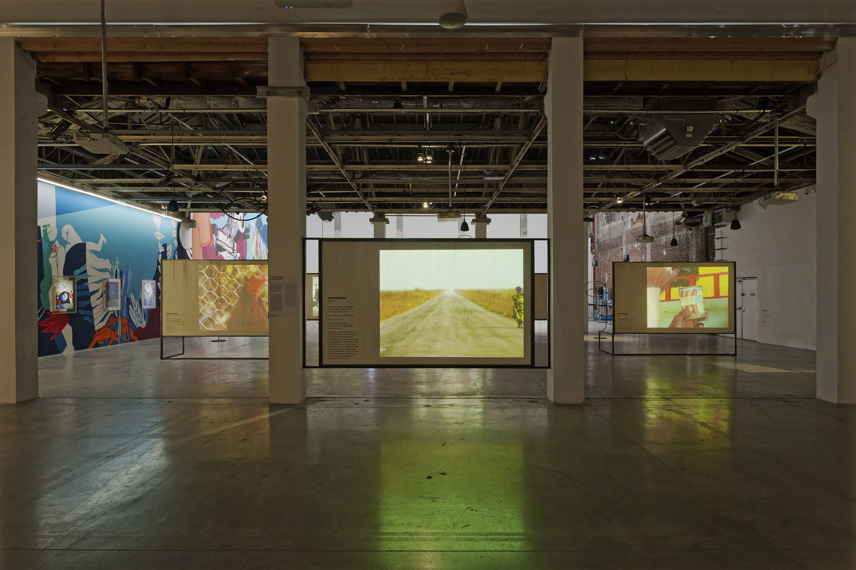 Installation view with films projected onto rectangular apparatuses. A colorful wall mural to the left extends backwards. Ceiling scaffolding is exposed.