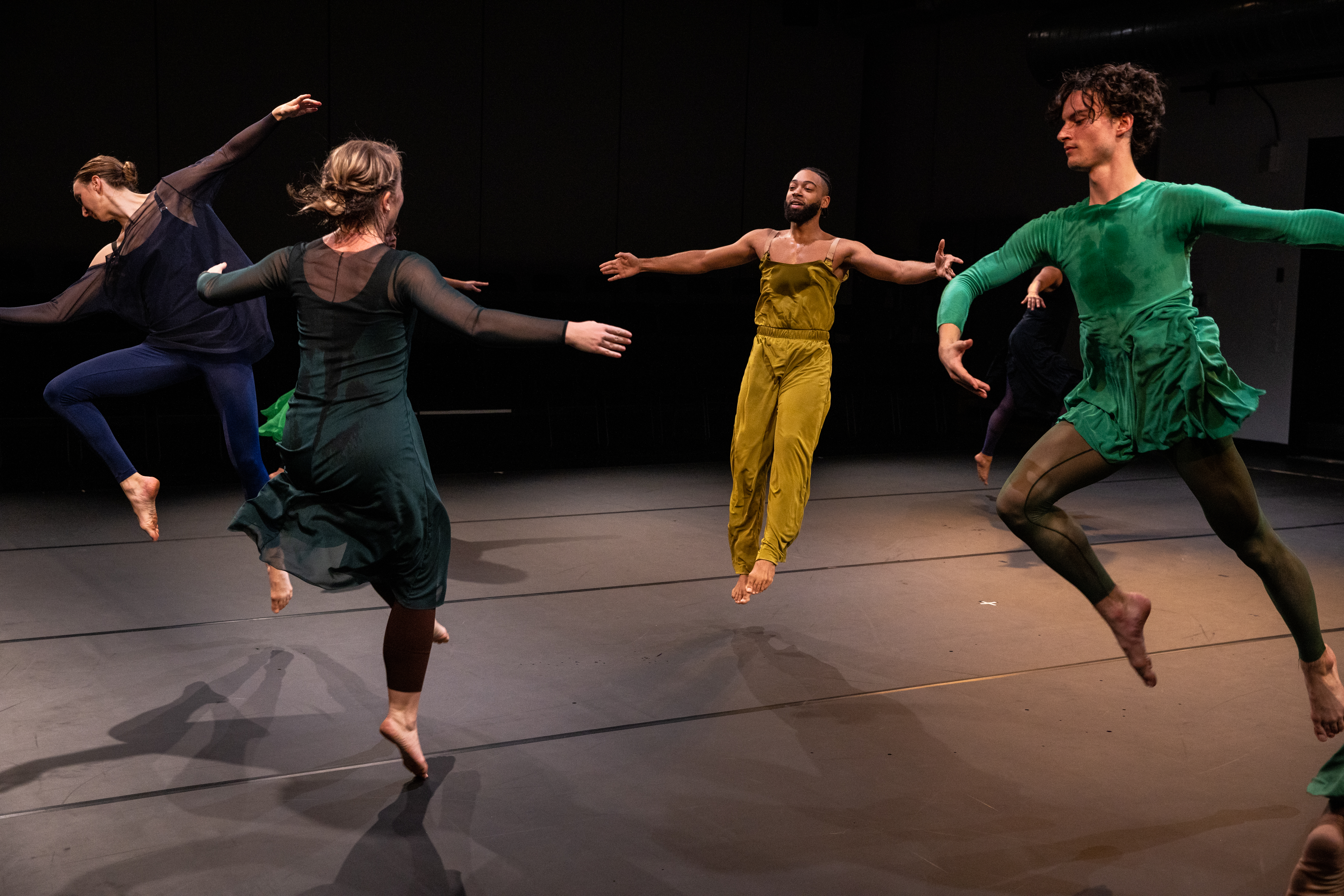 Four dancers in earth-toned, flowing outfits are leaping in air in a circular formation.