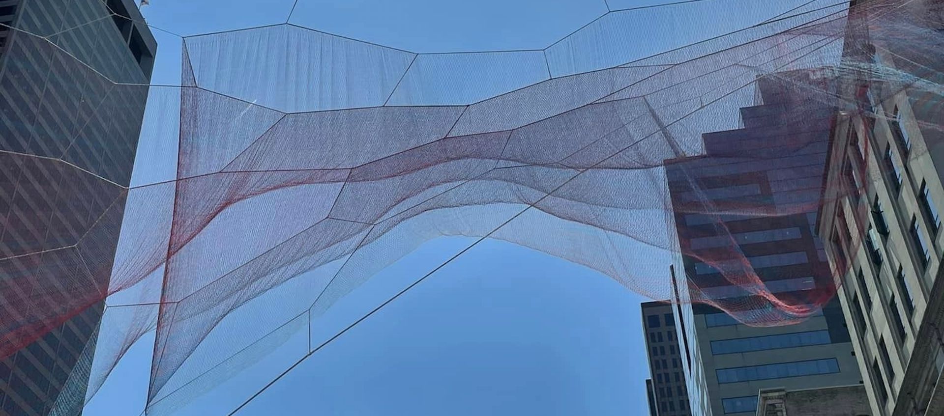 A blue sky is bordered by buildings. A large, translucent sculpture made of red netting is suspended in the air between the buildings.